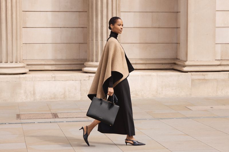 Elegant woman in cape and black dress walks confidently, holding a black leather tote bag in an urban setting with architectural details.