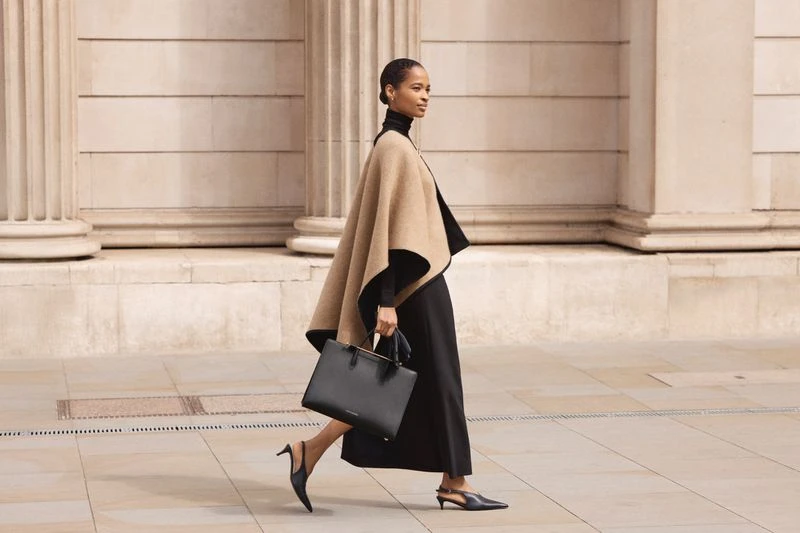 Elegant woman in cape and black dress walks confidently, holding a black leather tote bag in an urban setting with architectural details.