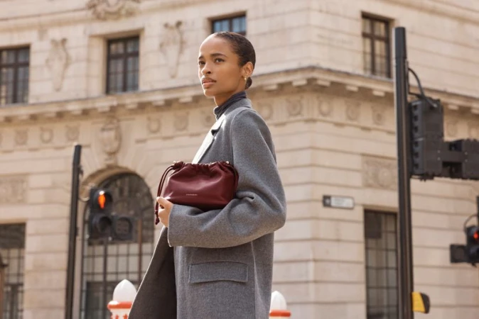 Model Ana Flávia wearing an oversized grey wool coat and holding a burgundy leather pouch bag on a city street.