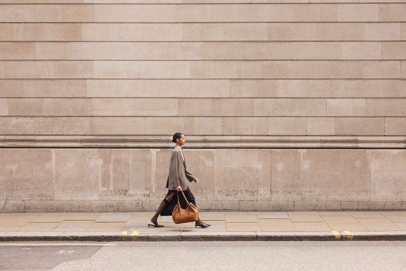 A woman in a grey blazer and black skirt walks on a sidewalk next to a building with tan stone walls, carrying a brown leather bag.