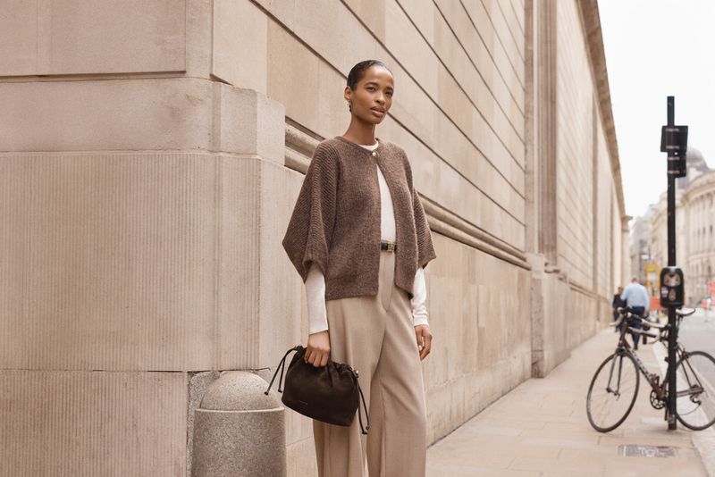 Woman in brown cape-style sweater, beige trousers, and black purse stands on city sidewalk with bike in the background.