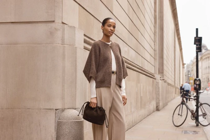 Woman in brown cape-style sweater, beige trousers, and black purse stands on city sidewalk with bike in the background.