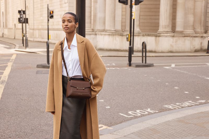 Elegant woman in tan coat with brown leather handbag crosses city street in daytime, featuring a modern fashion aesthetic and urban setting.
