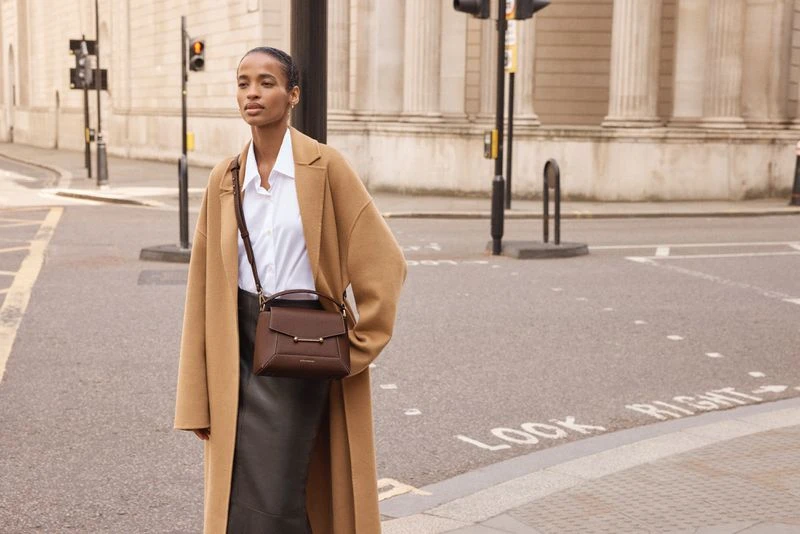 Elegant woman in tan coat with brown leather handbag crosses city street in daytime, featuring a modern fashion aesthetic and urban setting.