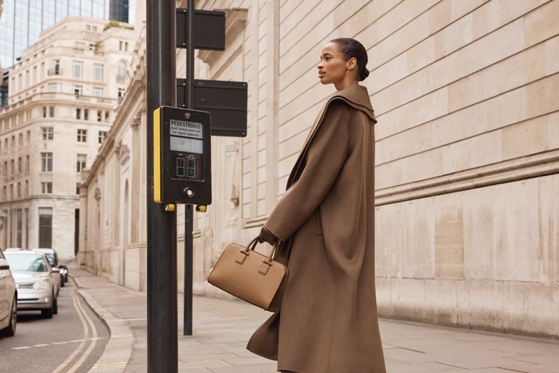A fashionable woman in a brown coat walks on a city street carrying a tan purse, showcasing urban style with a pedestrian crossing signal nearby.