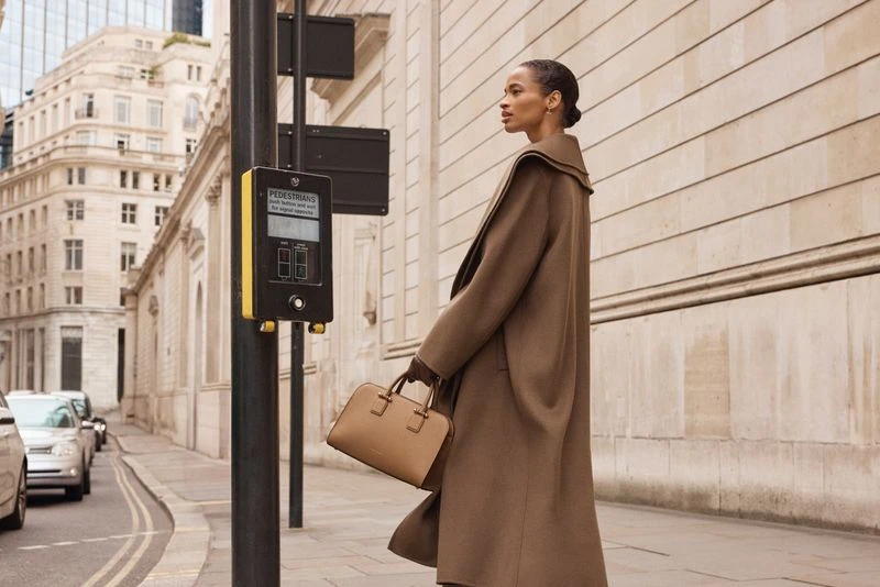 A fashionable woman in a brown coat walks on a city street carrying a tan purse, showcasing urban style with a pedestrian crossing signal nearby.