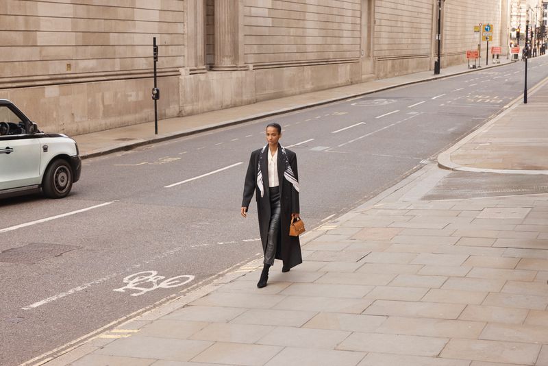 A stylish woman in a black coat strides down an urban street, carrying a brown handbag, with a London taxi cab in the background.