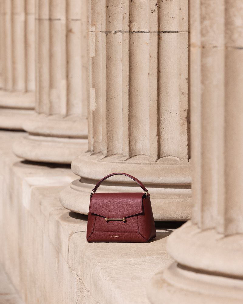 Burgundy handbag displayed in front of the stone pillar, with soft, natural light highlighting its texture and classic design.