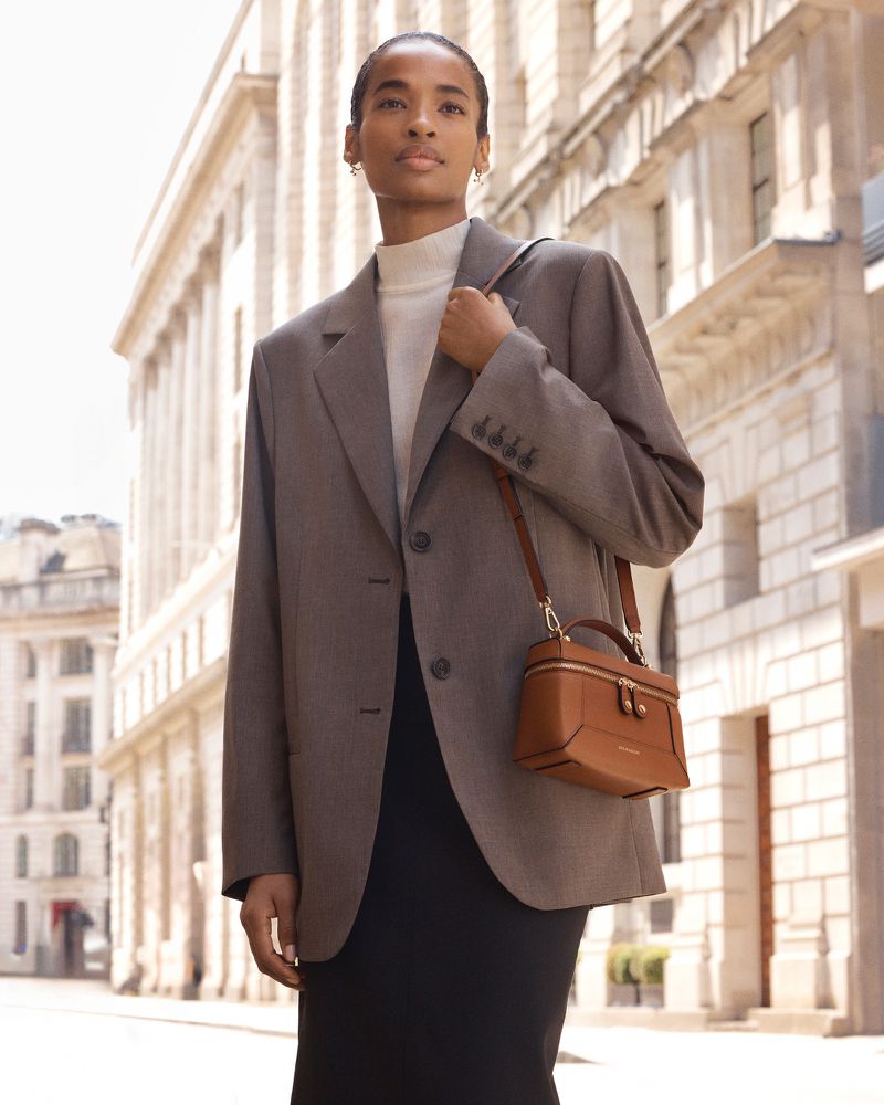 Model Ana Flávia wearing an oversized grey blazer and carrying a tan leather vanity bag along a city street.