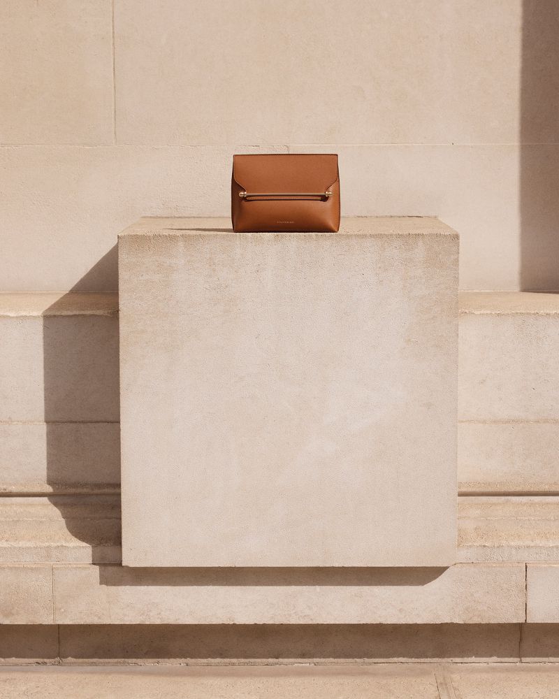 Tan-colored leather handbag with a metal bar detail is centrally positioned on a rectangular block against a light tan neutral wall background.