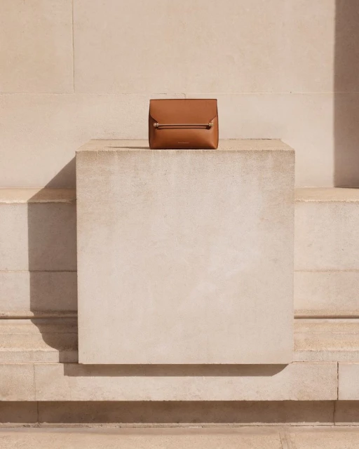 Tan-colored leather handbag with a metal bar detail is centrally positioned on a rectangular block against a light tan neutral wall background.