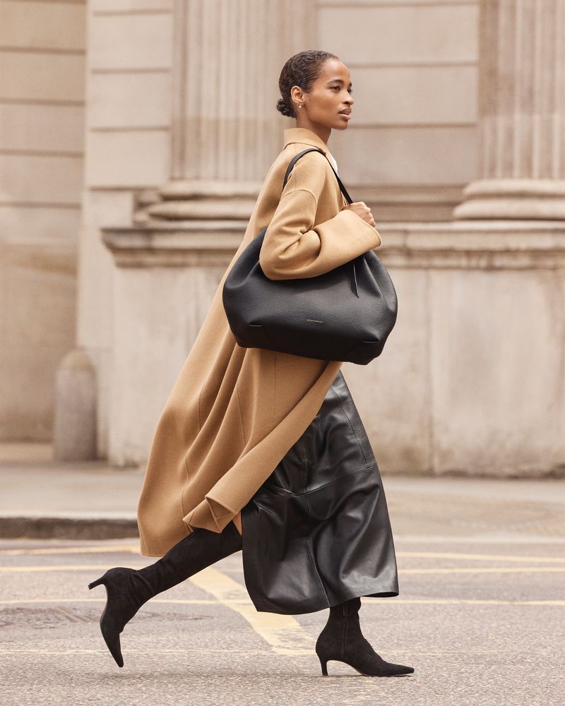 Model Ana Flávia in a long camel coat carrying a large black leather shoulder bag while walking along a city street.