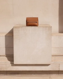 Brown leather handbag with a metal bar closure resting on a square stone plinth in direct sunlight.
