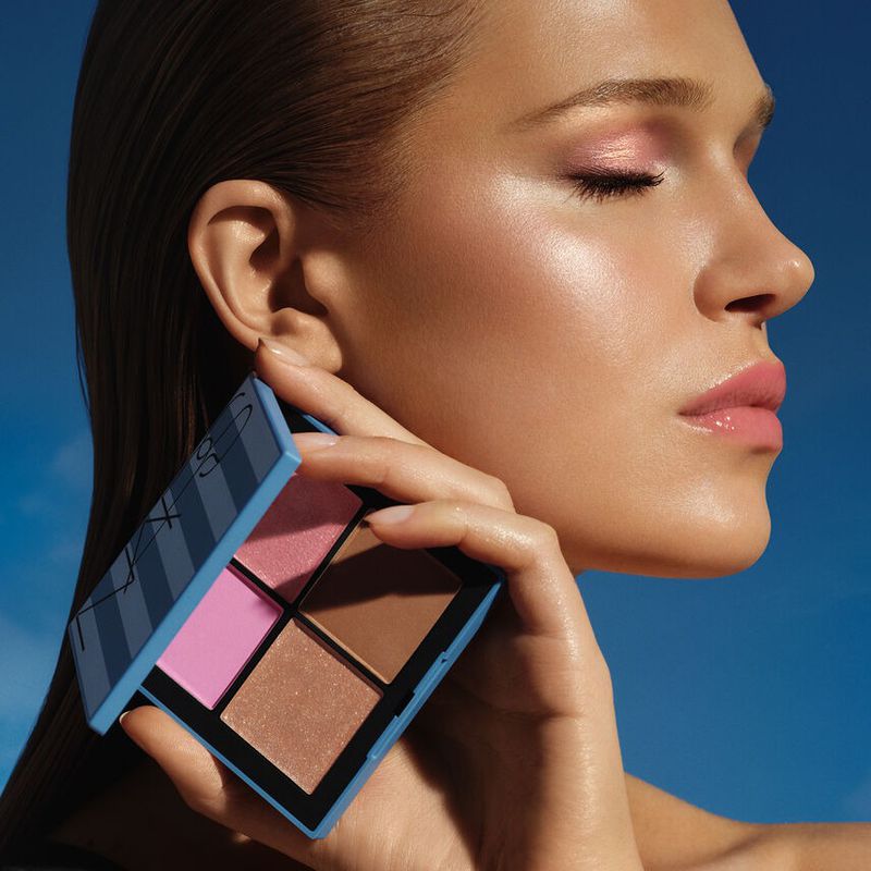 Close-up shot of fair-skinned woman holding an open NARS makeup palette with various shades against a blurred blue background.