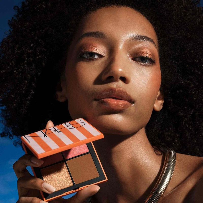 Woman holding NARS orange striped makeup palette for beauty product shot with bronze skin, dark curly hair and a silver choker necklace.
