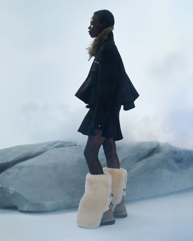 Fashion shot of a Black woman in a dark jacket and skirt with furry boots in a studio with diffused lighting and a boulder.