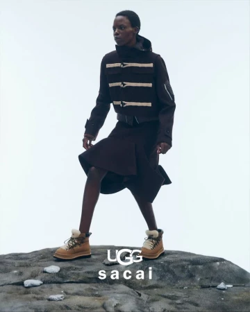 Fashion shot of a Black female model in a brown coat and skirt standing on a rock, wearing UGG boots. UGG x sacai logo is visible.