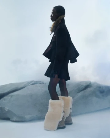 Agel Akol poses in a black flared jacket, dark miniskirt, and oversized cream shearling boots against a snowy studio set.