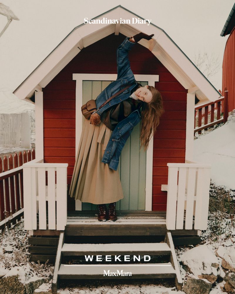 Female model posing in front of a small red house, wearing a denim jacket, tan skirt, and red boots in a snowy, outdoor environment.