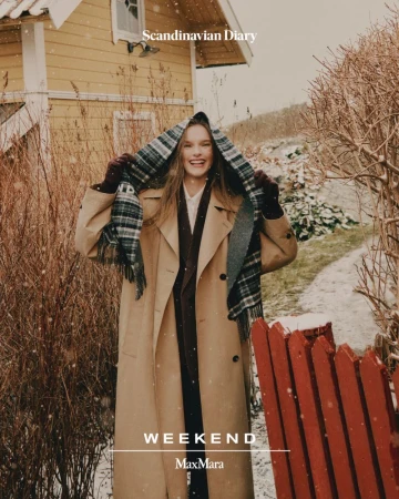 A smiling woman in a trench coat stands outdoors in a snowy setting, wearing a scarf and gloves with a red fence and house visible.