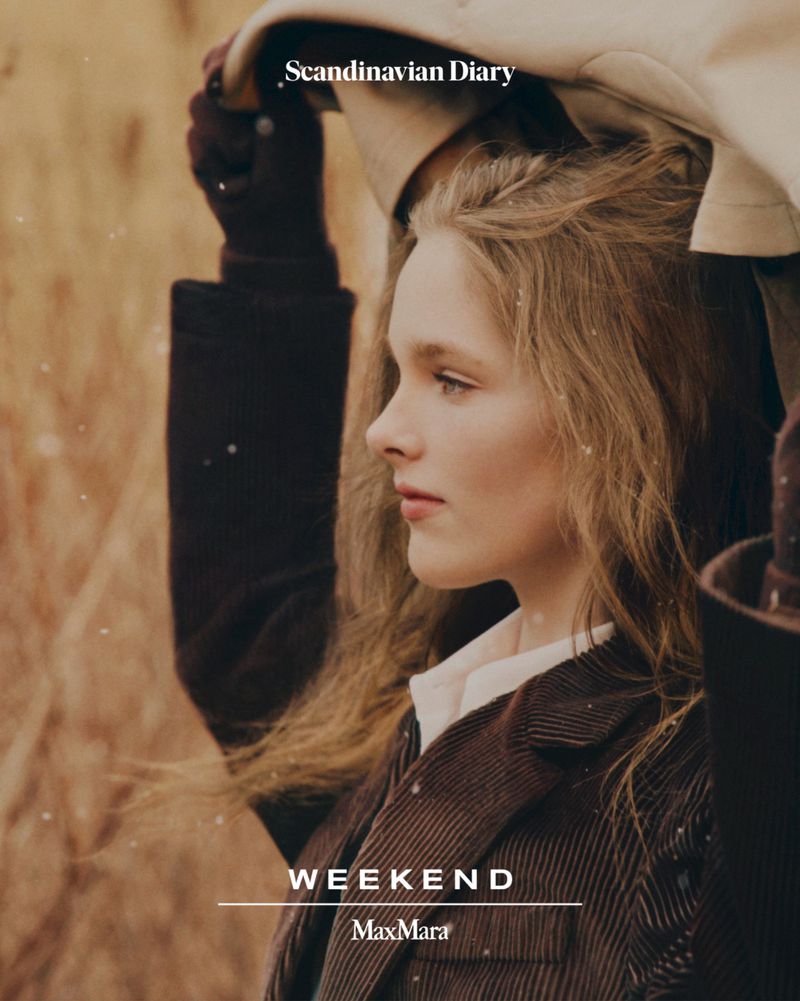 A young adult woman with wavy, blonde hair in a brown jacket poses in a field setting, lit by soft light, with visible fashion branding.