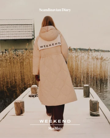 Woman in long puffer coat walks along a snow-covered dock next to a body of water, featuring the text "Weekend" and brand name Max Mara.