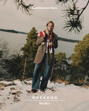 Model Vanessa Warg wearing a grey tweed blazer, wide-leg denim jeans, and a striped scarf on snowy terrain overlooking water.