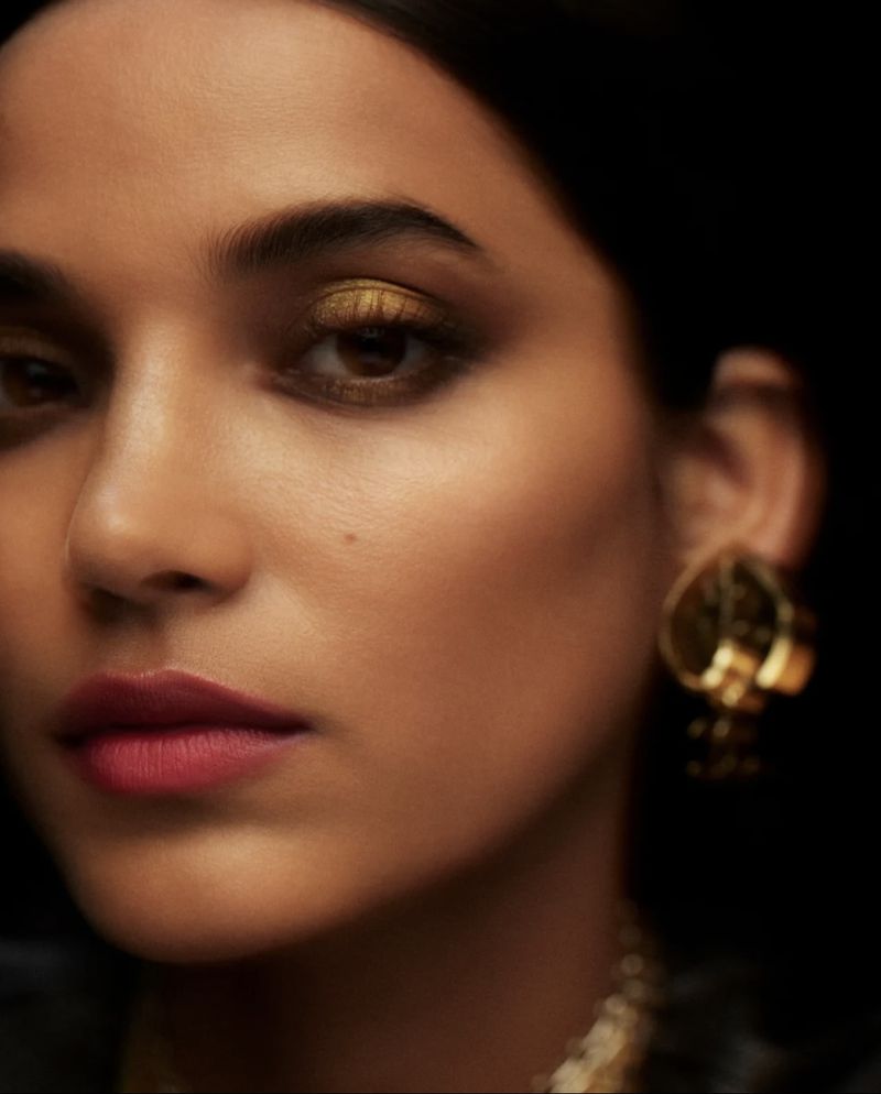 Close-up portrait of a young woman with golden eyeshadow, pink lipstick, and gold jewelry against a dark background in a studio setting.
