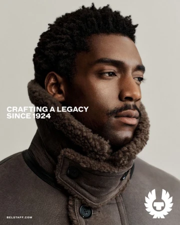 Close-up studio shot of a dark-skinned adult male model wearing a brown shearling-lined Belstaff coat in a fashion advertisement.