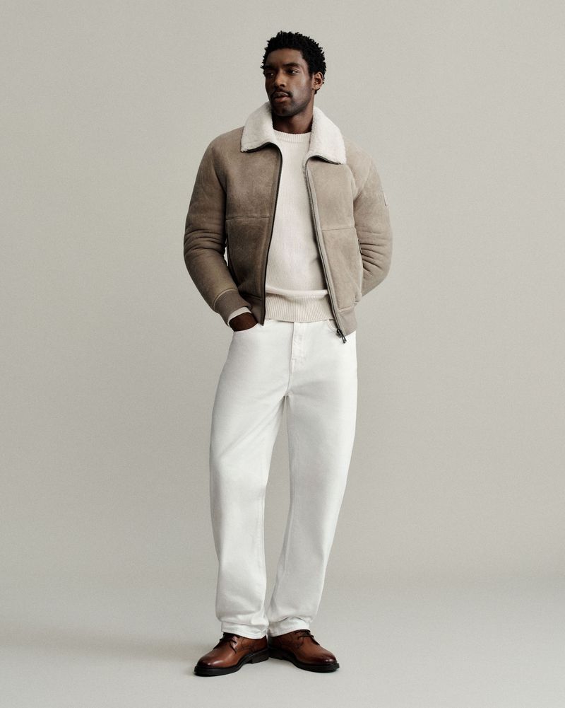 Studio shot of a Black male model wearing a beige shearling bomber jacket, cream sweater, white jeans, and brown leather shoes.