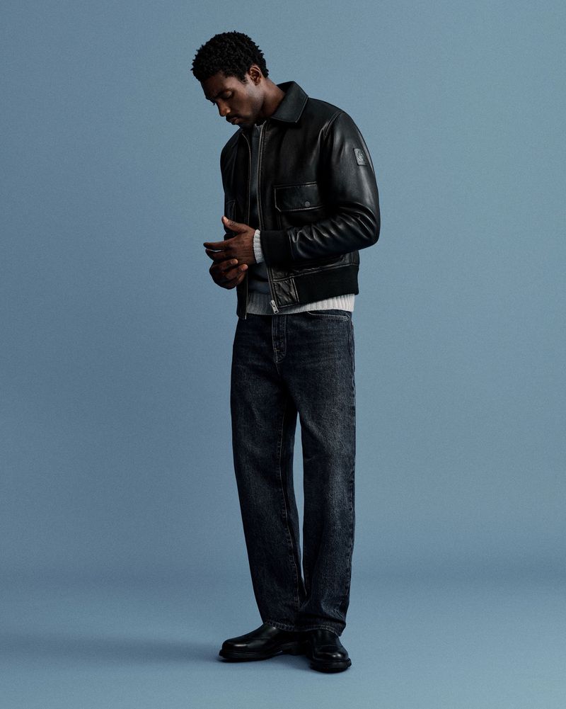 Man in a black leather jacket and dark wash jeans stands against a blue background in a fashion shot with studio lighting.