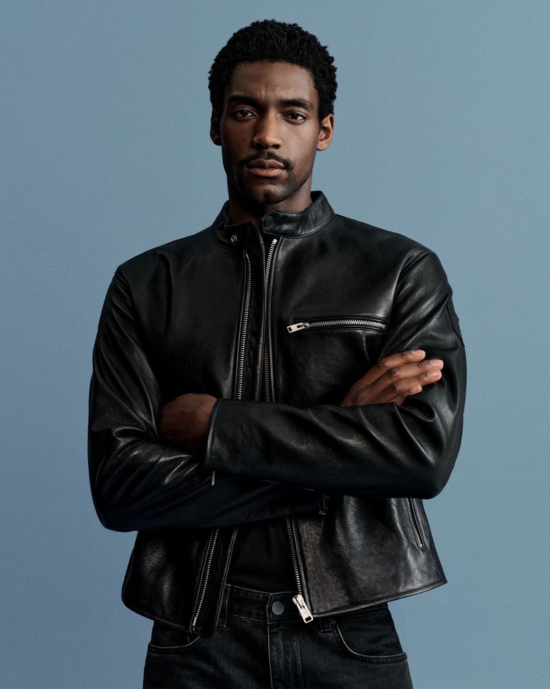 A confident Black man with a mustache poses indoors, wearing a black leather jacket, jeans, and a black shirt against a blue background.