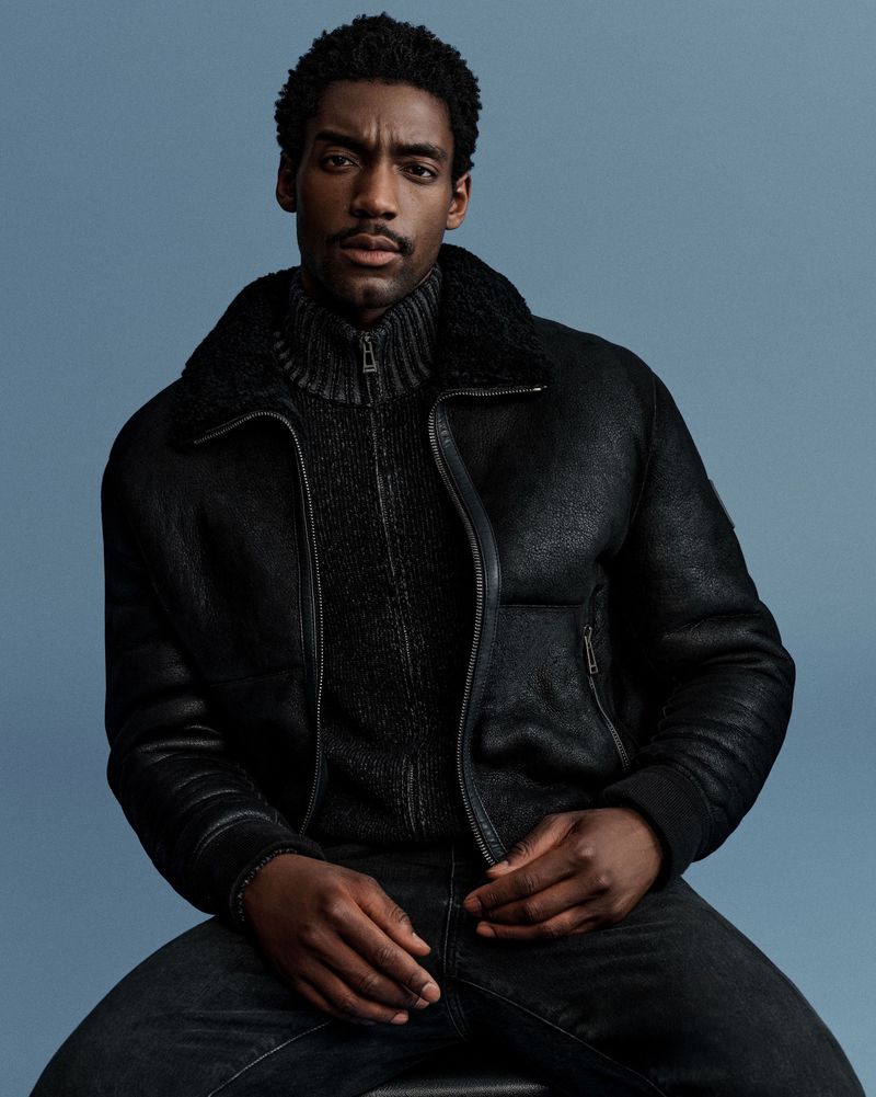 Portrait of a Black male model wearing a black jacket and jeans, seated against a muted blue background in a photo studio.