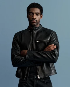 Model Halid Nuhu posing with arms crossed in a black leather moto jacket against a solid light blue studio backdrop.