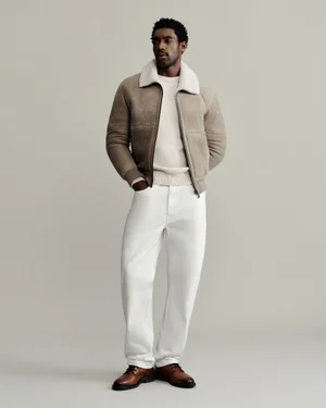 Halid Nuhu wearing a beige shearling bomber jacket, cream sweater, white trousers, and brown leather shoes in a studio setting.