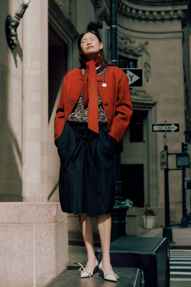 Asian woman in a red jacket and navy skirt poses on a city bench with architectural buildings and a street sign in the background.