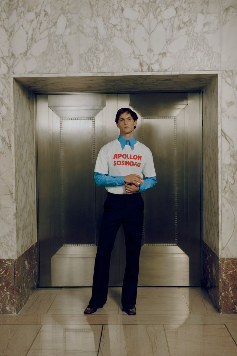 Stylish man poses in front of elevator wearing a graphic t-shirt, blue collared shirt, and dark pants inside a building with marble walls.