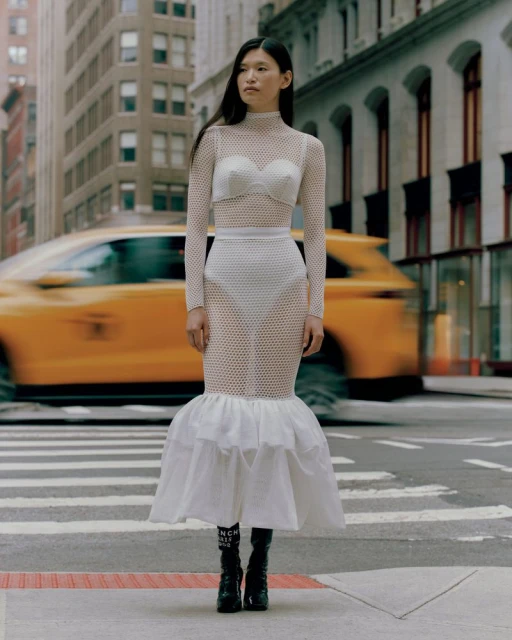 Woman in a see-through dress stands on a city crosswalk, with a blurred yellow taxi and urban buildings in the background.