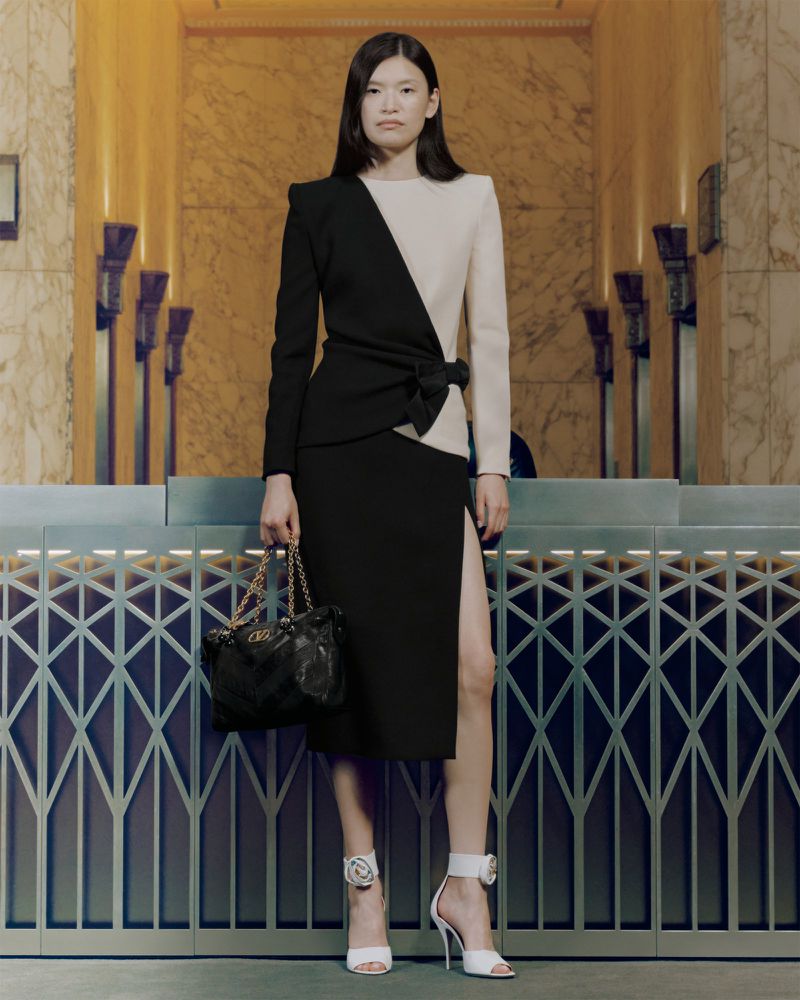 Elegant Asian woman in a split-tone black and beige dress, holding a purse, and posing in white heeled sandals against a classic background.