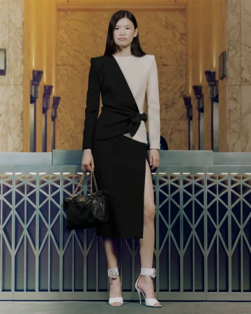 Elegant Asian woman in a split-tone black and beige dress, holding a purse, and posing in white heeled sandals against a classic background.