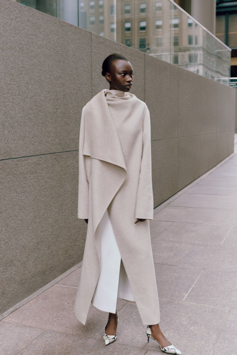 A full-length portrait showcases a slender Black woman in a beige coat, white pants, and heels, posing against an urban backdrop of neutral tones.
