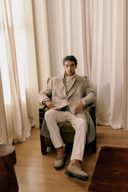 Elegant adult male seated in an armchair, wearing a coat, turtleneck sweater, and white pants, complemented by light brown boots, indoors.