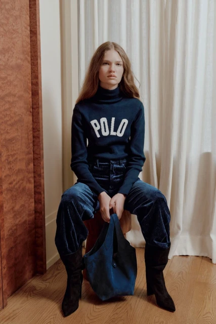 Woman in a blue turtleneck sweater with the word POLO on it, paired with velvet pants and black boots, sitting indoors and holding a handbag.