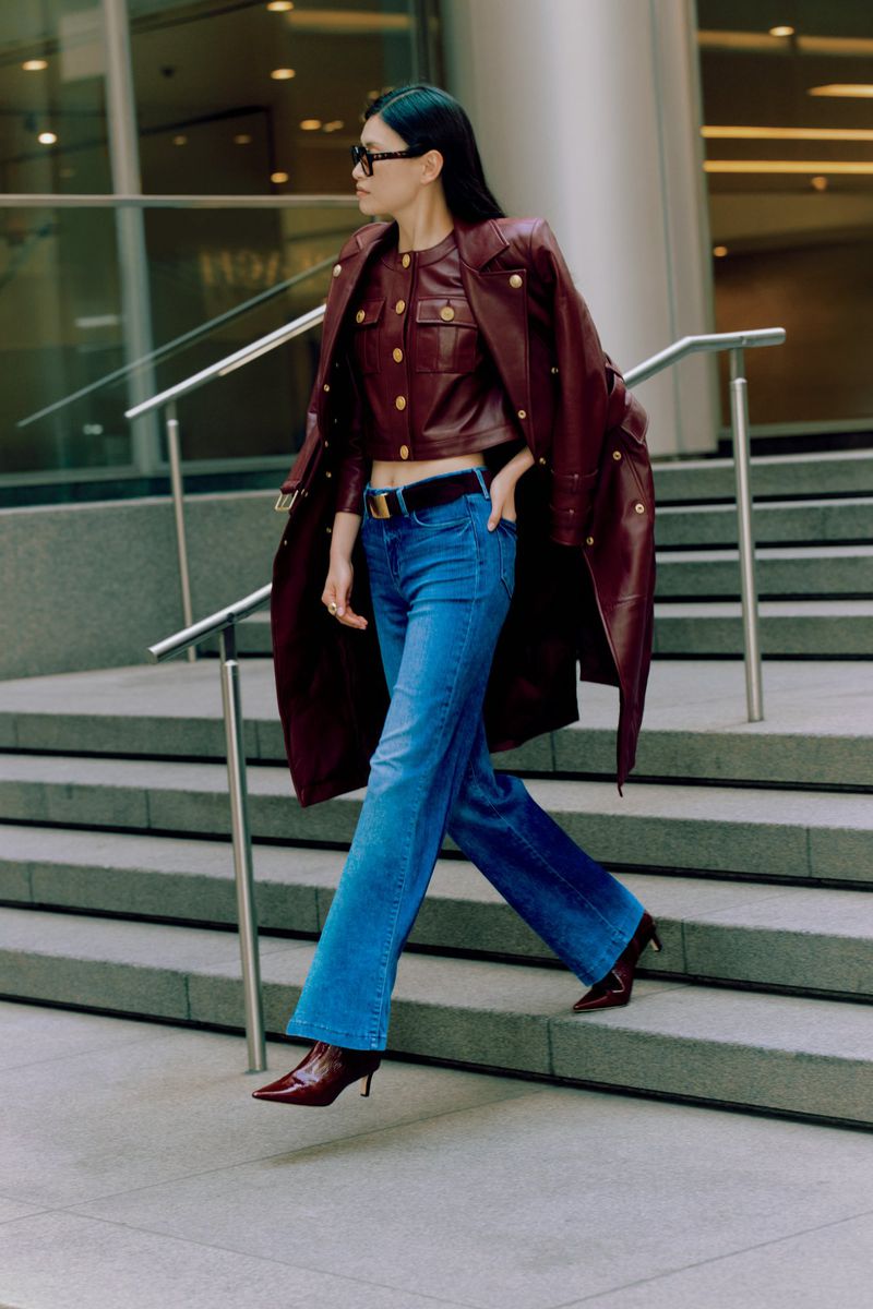 Woman in burgundy leather coat and jeans walking on concrete stairs, accessorized with sunglasses and a black belt, urban setting.
