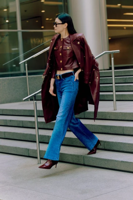 Woman in burgundy leather coat and jeans walking on concrete stairs, accessorized with sunglasses and a black belt, urban setting.