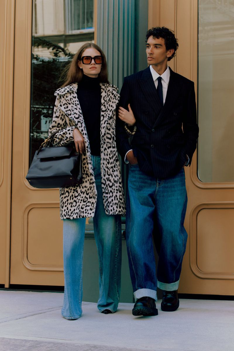 Stylish couple in a fashion editorial shot, wearing a leopard print coat and a navy suit, standing in front of a glass door.