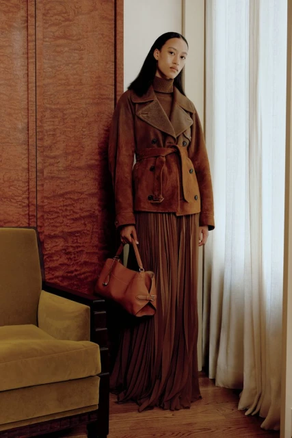 A woman poses in a brown belted jacket and long pleated skirt, holding a handbag, in an indoor setting with neutral tones and soft lighting.