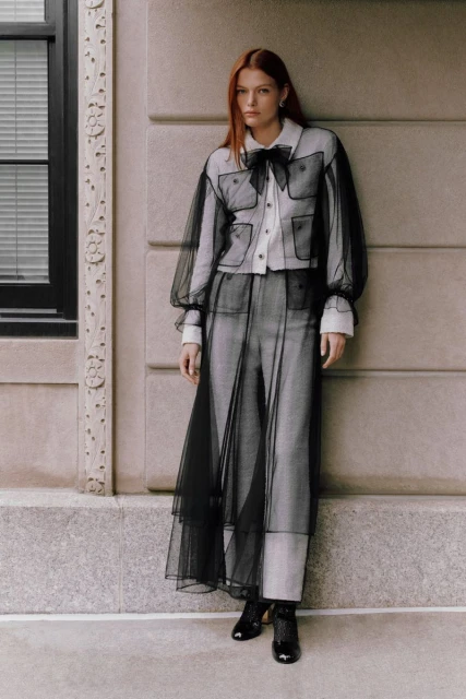 A redhead woman models neutral-toned fashion with black trim and tulle accents, standing in an urban setting with architectural details.