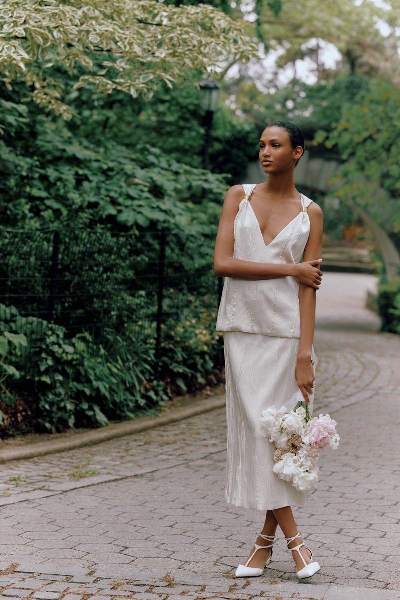 Woman in a cream two-piece dress stands on a brick path in a park, holding a bouquet of flowers, with lush green trees behind her.