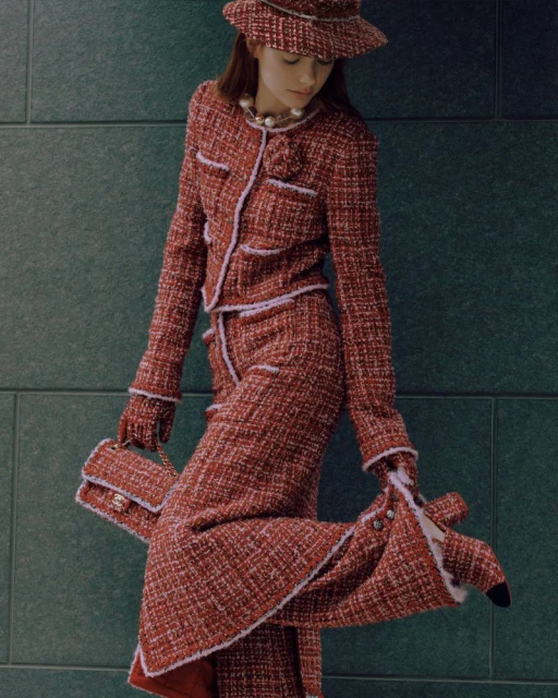 A stylish young woman in a red tweed suit and matching bucket hat poses against a textured gray wall, holding a small red tweed handbag.